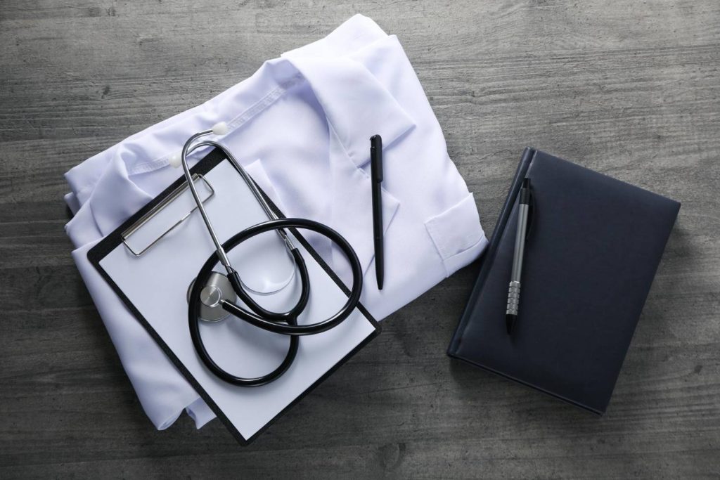 Bata médica blanca doblada con un estetoscopio y un portapapeles encima, junto a una libreta oscura sobre una mesa de madera gris; concepto visual para casos de negligencia médica en Chile.