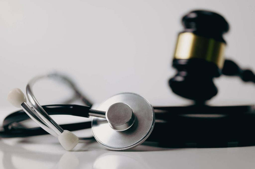 Primer plano de un estetoscopio médico negro sobre una superficie blanca, con un mazo de justicia desenfocado al fondo, conceptualizando demandar por negligencia médica.