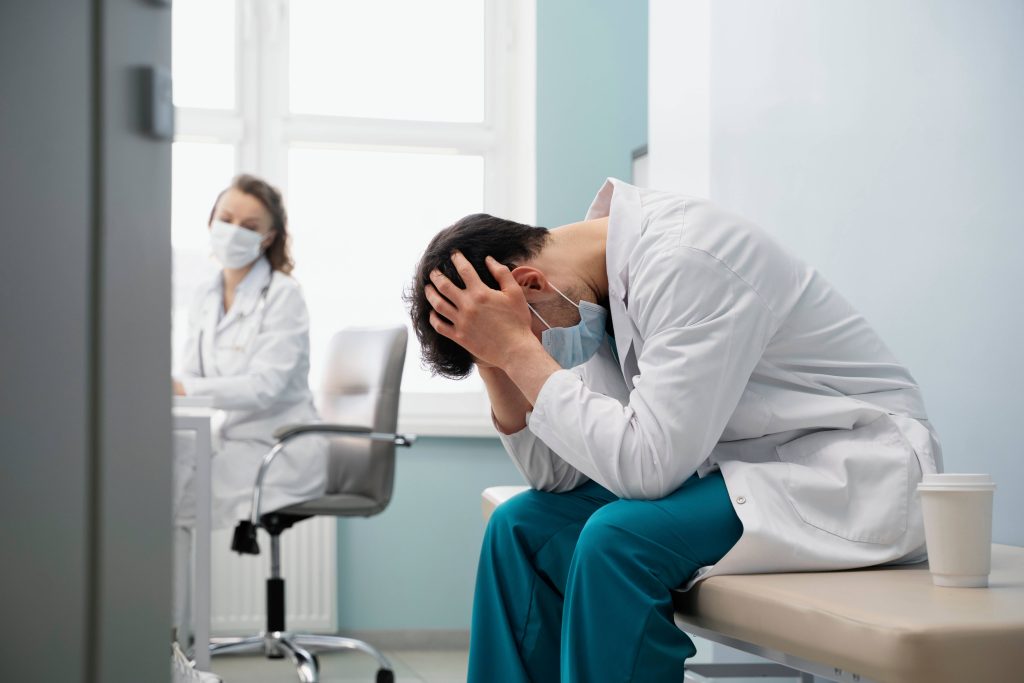 Médico angustiado con la cabeza entre las manos, sentado en un banco de examen con una colega borrosa al fondo de una sala de hospital; una escena que ilustra el estrés asociado a situaciones que requieren mediación por negligencia médica en Chile.