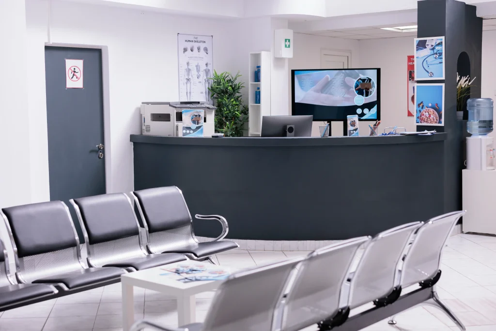 Un plano de gran angular de la sala de espera de una clínica moderna y vacía, con filas de sillas de espera de metal y un mostrador de recepción curvo y oscuro. En el mostrador de recepción hay equipos de oficina y una gran pantalla de TV, y en la pared hay carteles médicos y anatómicos. Esta imagen representa el área de atención al paciente, donde el personal debe recordar que es obligatorio para hospitales y clínicas entregar la ficha clínica a los pacientes que la soliciten.
