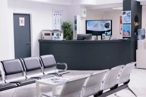 Un plano de gran angular de la sala de espera de una clínica moderna y vacía, con filas de sillas de espera de metal y un mostrador de recepción curvo y oscuro. En el mostrador de recepción hay equipos de oficina y una gran pantalla de TV, y en la pared hay carteles médicos y anatómicos. Esta imagen representa el área de atención al paciente, donde el personal debe recordar que es obligatorio para hospitales y clínicas entregar la ficha clínica a los pacientes que la soliciten.